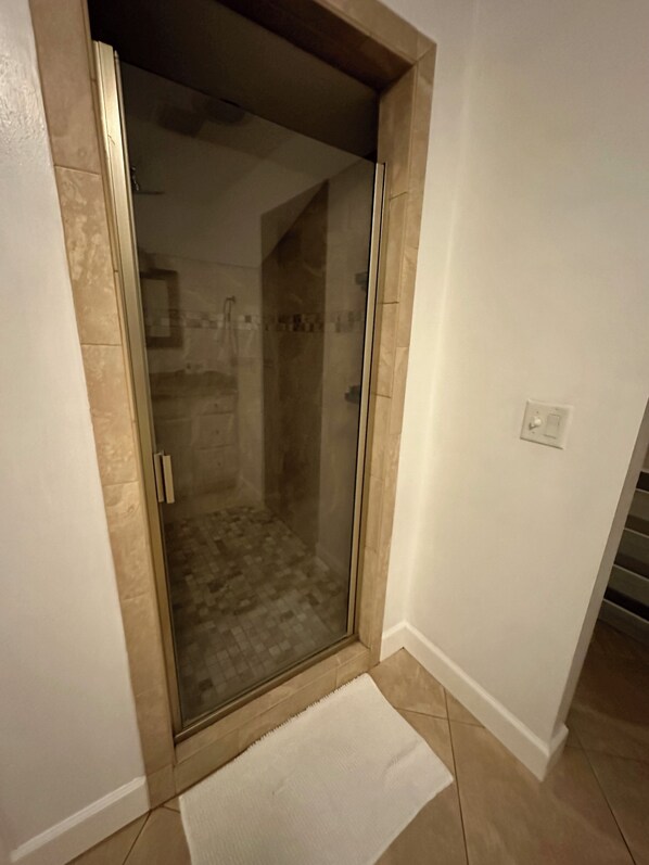 Un área de ducha con paredes de vidrio, suelo de baldosas, adyacente a una pared y una puerta blancos.