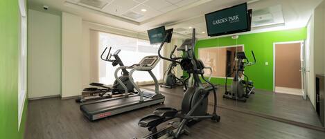 Un gimnasio con cintas de correr, máquinas elípticas y una pantalla mostrando 'GARDEN PARK HOTELS'.