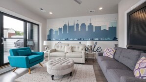 Una sala moderna con un mural de un horizonte urbano, un sillón verde azulado, un sofá gris y un reposapiés blanco.