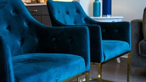 Una habitación con dos sillones de terciopelo azul con patas doradas, una mesita auxiliar blanca con un jarrón azul y un sofá gris.