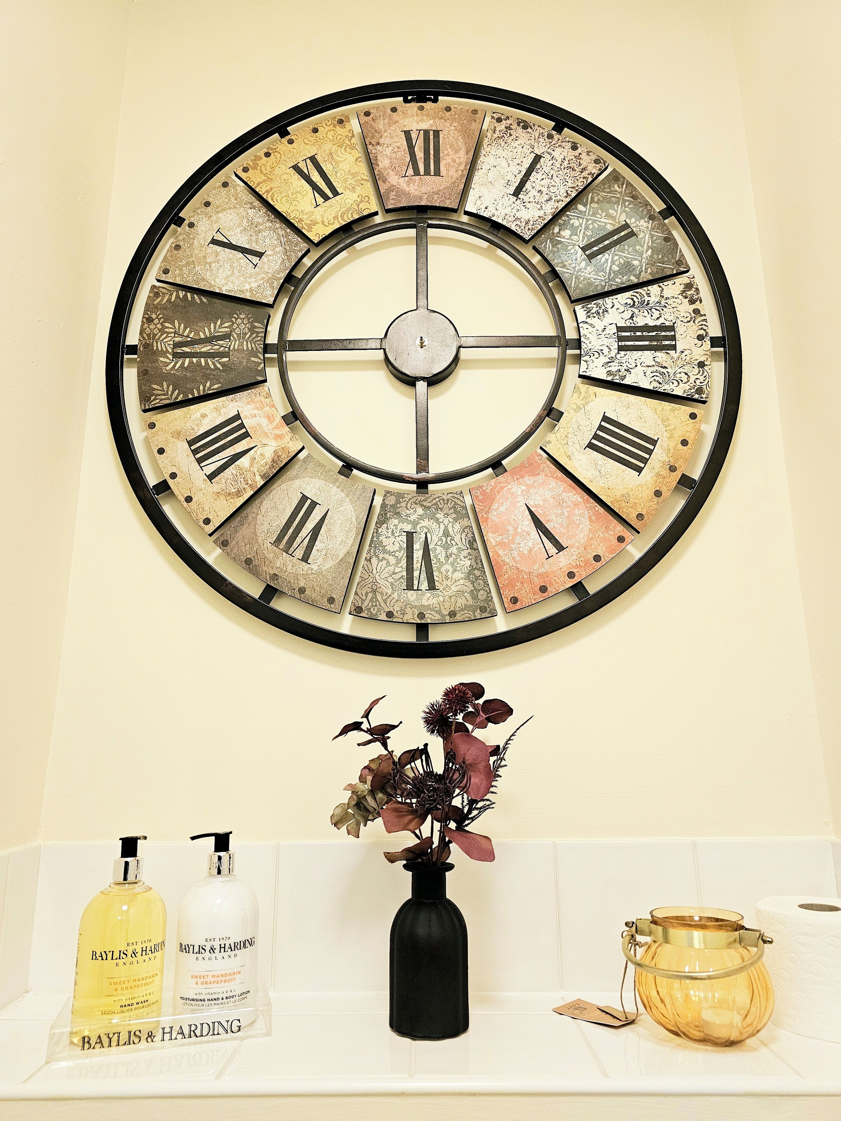 A wall clock with Roman numerals and a vase with dried flowers on a shelf.