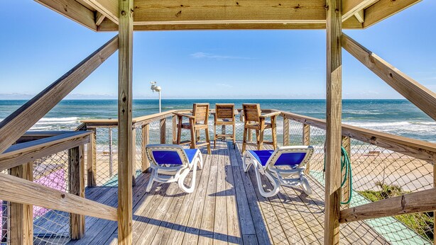 Eine Holzterrasse mit zwei Liegestühlen und einem Tisch, mit Blick auf einen Strand und eine klare Meeransicht.