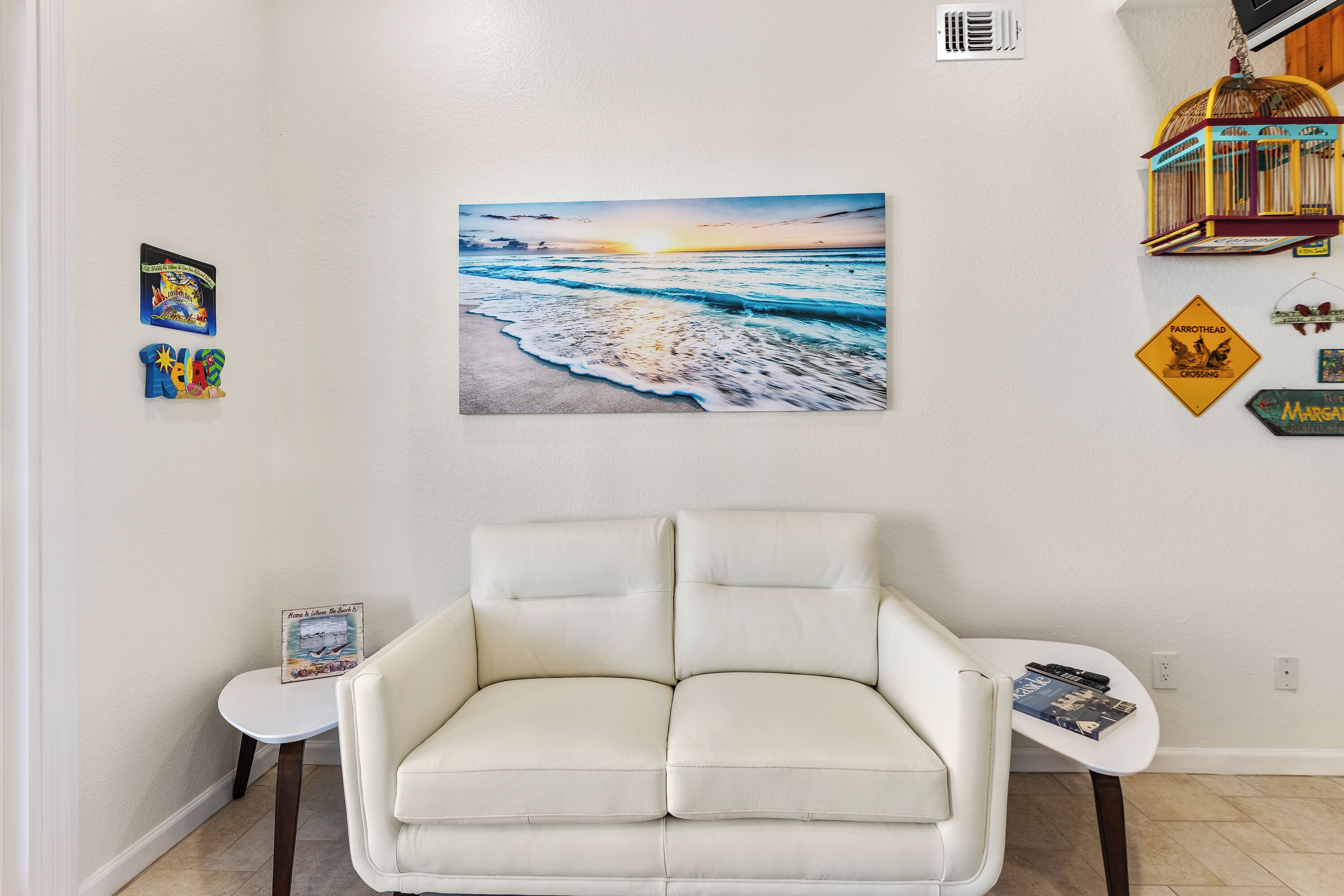 Een woonkamer met een witte bank, een klein witte tafeltje en een aan de muur hangend schilderij van een strandgezicht.