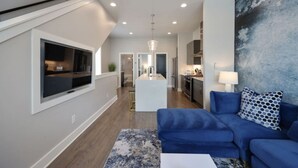 A modern living room with a blue sofa, a TV mounted on the wall, and a kitchen in the background. - West End Luxe | Rooftop Deck + Skyline (Nashville)