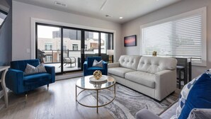 A modern living room with a blue sofa, a white sofa, a round coffee table, and a large window with blinds. - West End Luxe | Rooftop Deck + Skyline (Nashville)