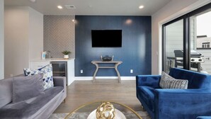 A modern living room with a grey sofa, a blue sofa, a flat-screen TV mounted on a dark wall, and a small table with a speaker. - West End Luxe | Rooftop Deck + Skyline (Nashville)