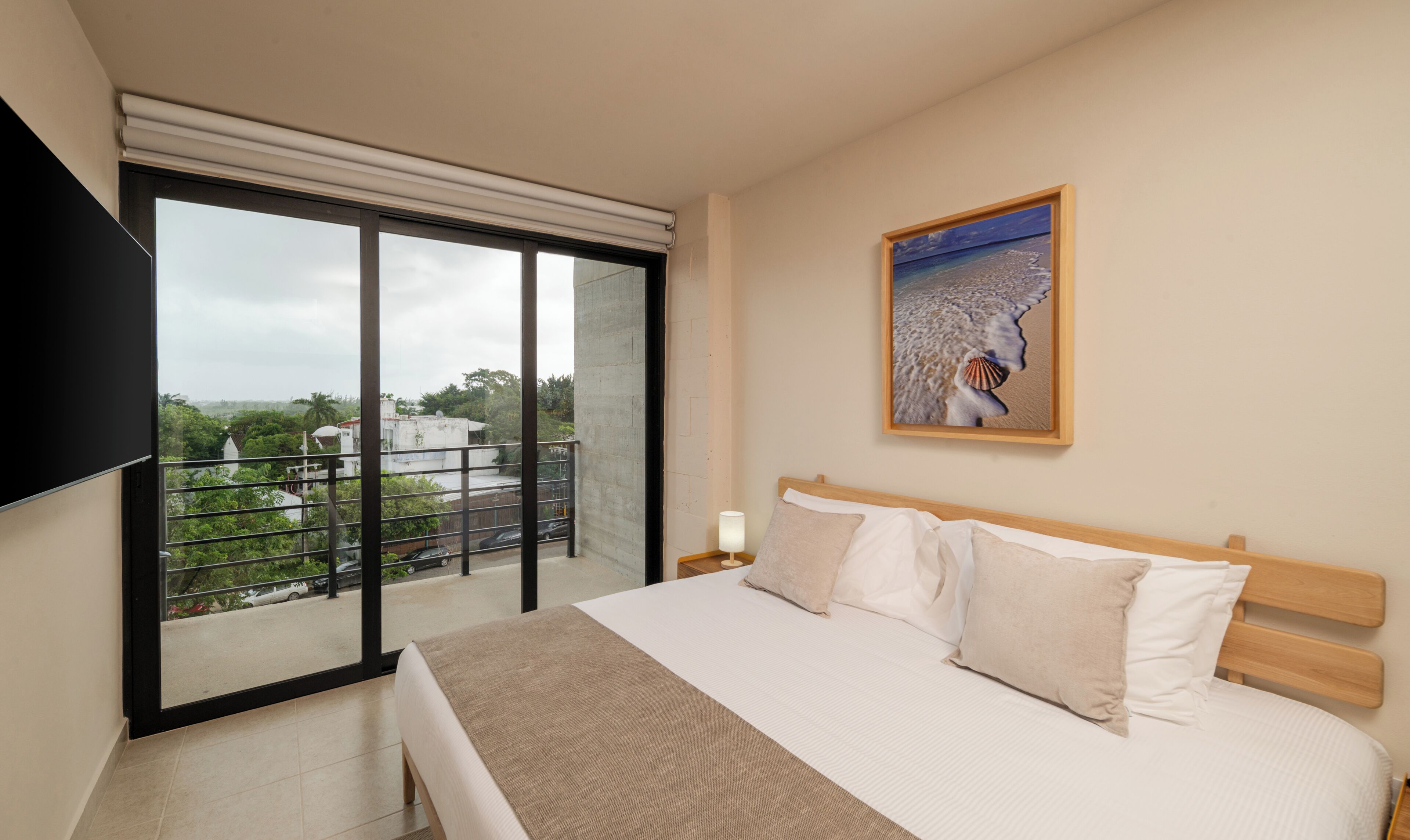 A hotel room with a bed, a nightstand, a wall-mounted TV, a painting, and a balcony with a view of buildings and trees.