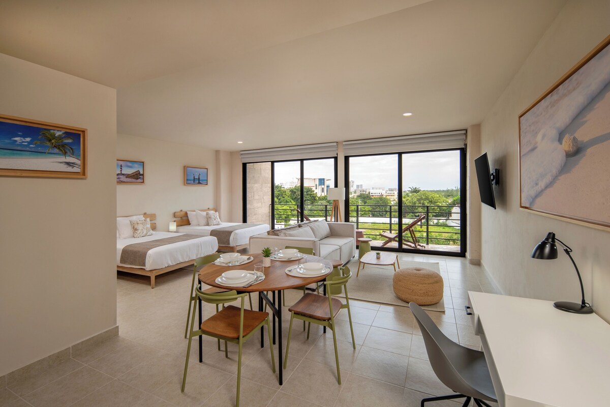 A modern hotel room with a bed, sofa, dining table, and chairs. There is a TV mounted on the wall and a window with a view of greenery.