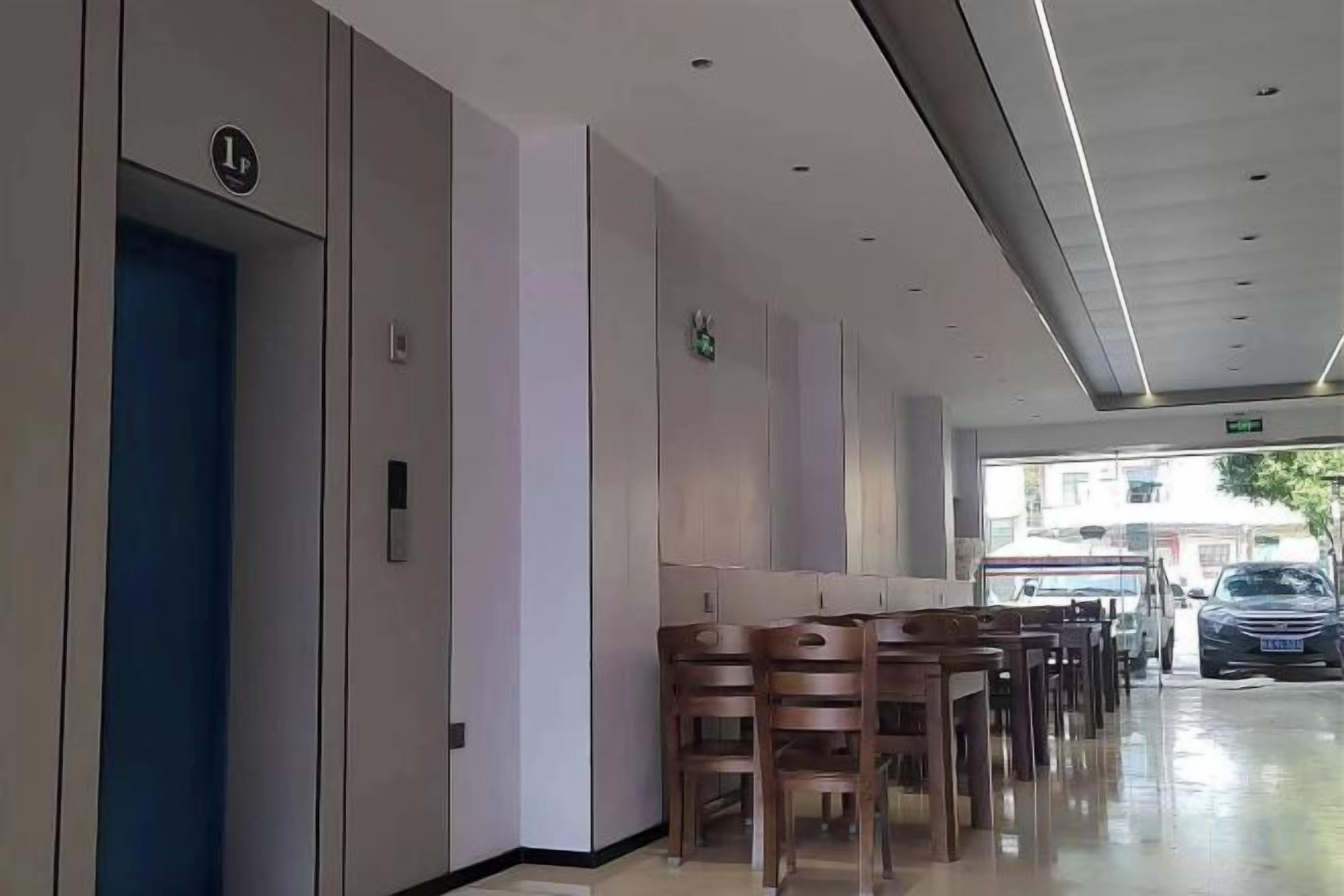 A hotel corridor with a blue elevator, a row of wooden chairs, and a car parked outside.