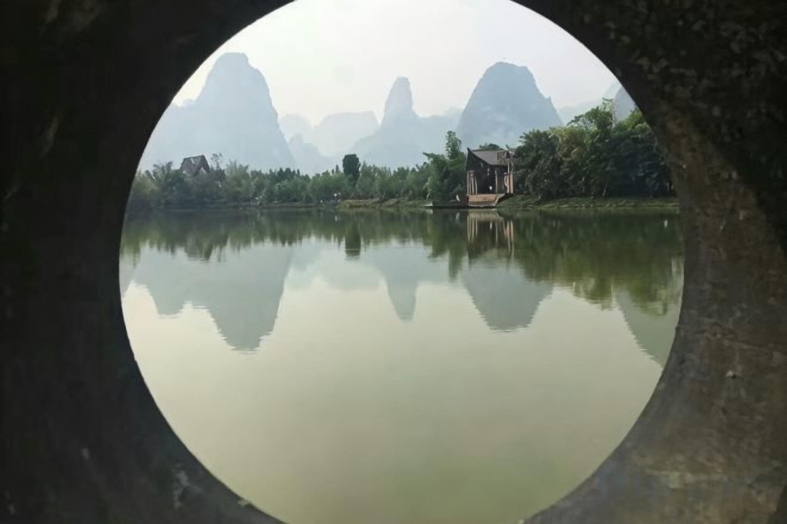 A circular view of a calm lake with a house on the shore, surrounded by mountains.