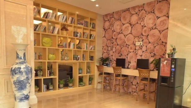 A reception area with a bookshelf, a large vase, a wall with a wood grain design, a desk with a computer, and a refrigerator. - GreenTree (Shuyang Development Zone Yingbin Avenue) (Shuyang)