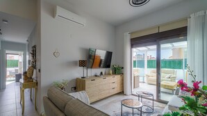 Ein modernes Wohnzimmer mit einer Couch, einem Holzschrank, einem Flachbildfernseher und einer Glastür, die zu einem Außenbereich mit Sitzgelegenheiten führt.