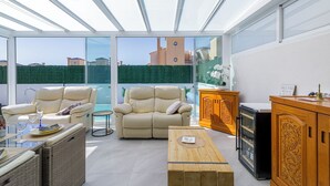 Ein sonnendurchfluteter Raum mit Glasdecke, einem beigefarbenen Sofa, einem Holztisch und einem kleinen Kühlschrank.