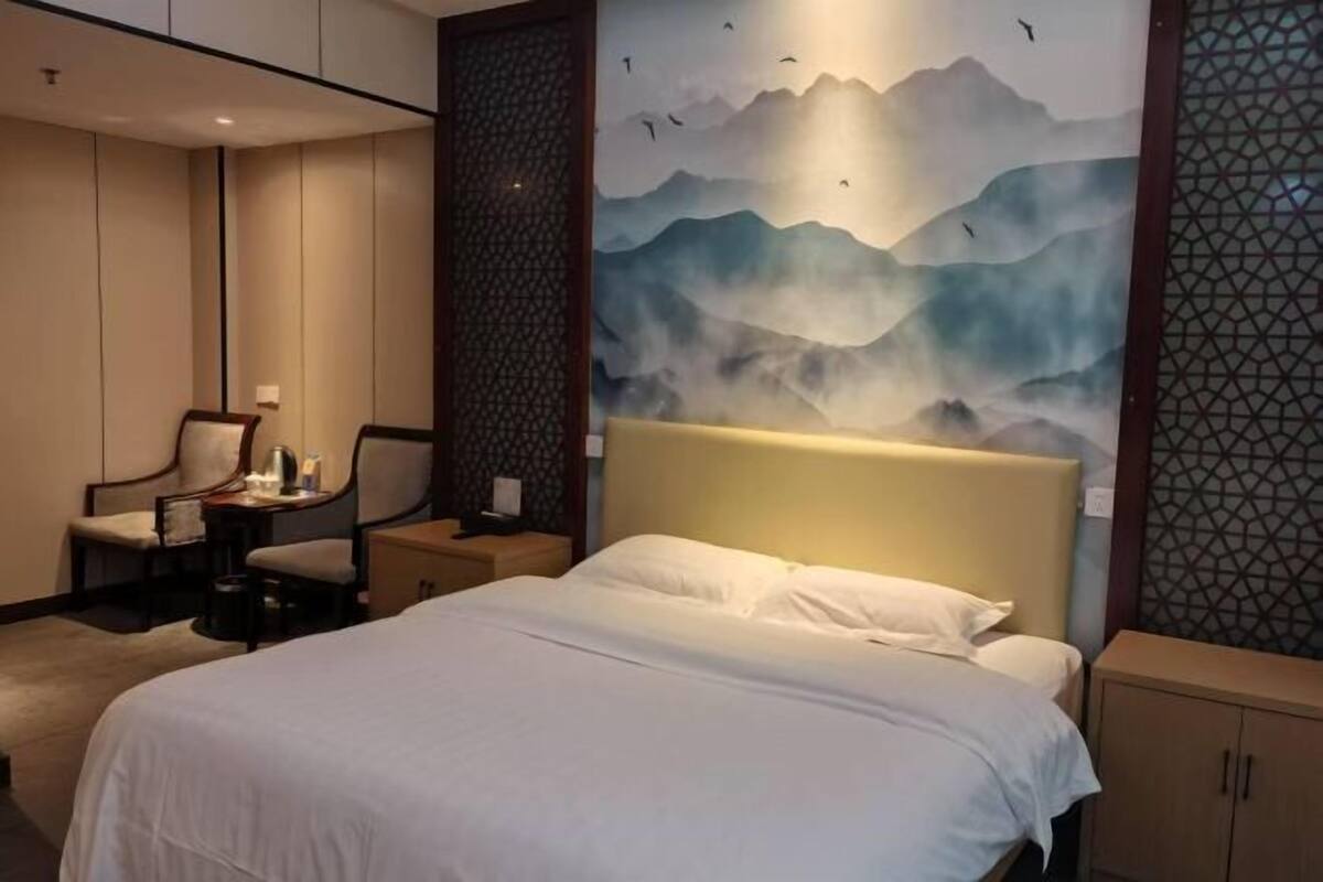 A hotel room with a bed, a desk with a chair, a nightstand, and a wall with a mountain painting.