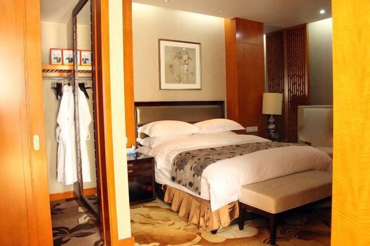 A hotel room with a bed, bedside table, lamp, and a wardrobe with hanging clothes.