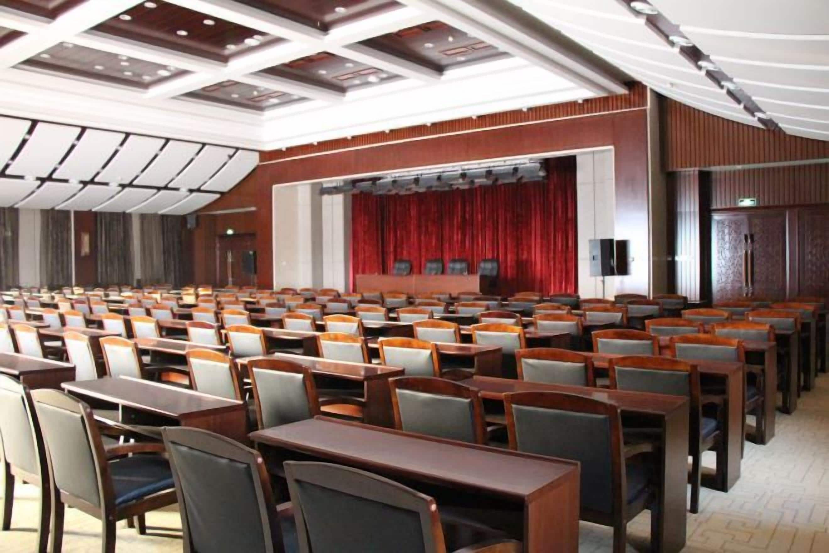 A conference room with rows of tables and chairs, a stage with a red curtain, and a ceiling with recessed lighting.