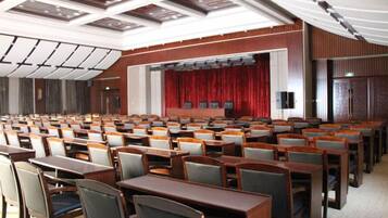 A conference room with rows of tables and chairs, a stage with a red curtain, and a ceiling with recessed lighting.