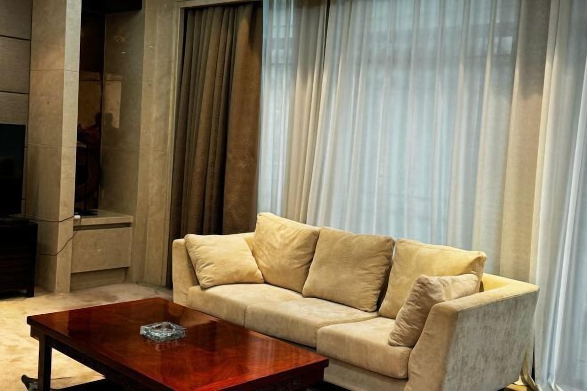 Una sala de estar con un sofá beige, una mesa de centro de madera y un ventanal con cortinas traslúcidas.