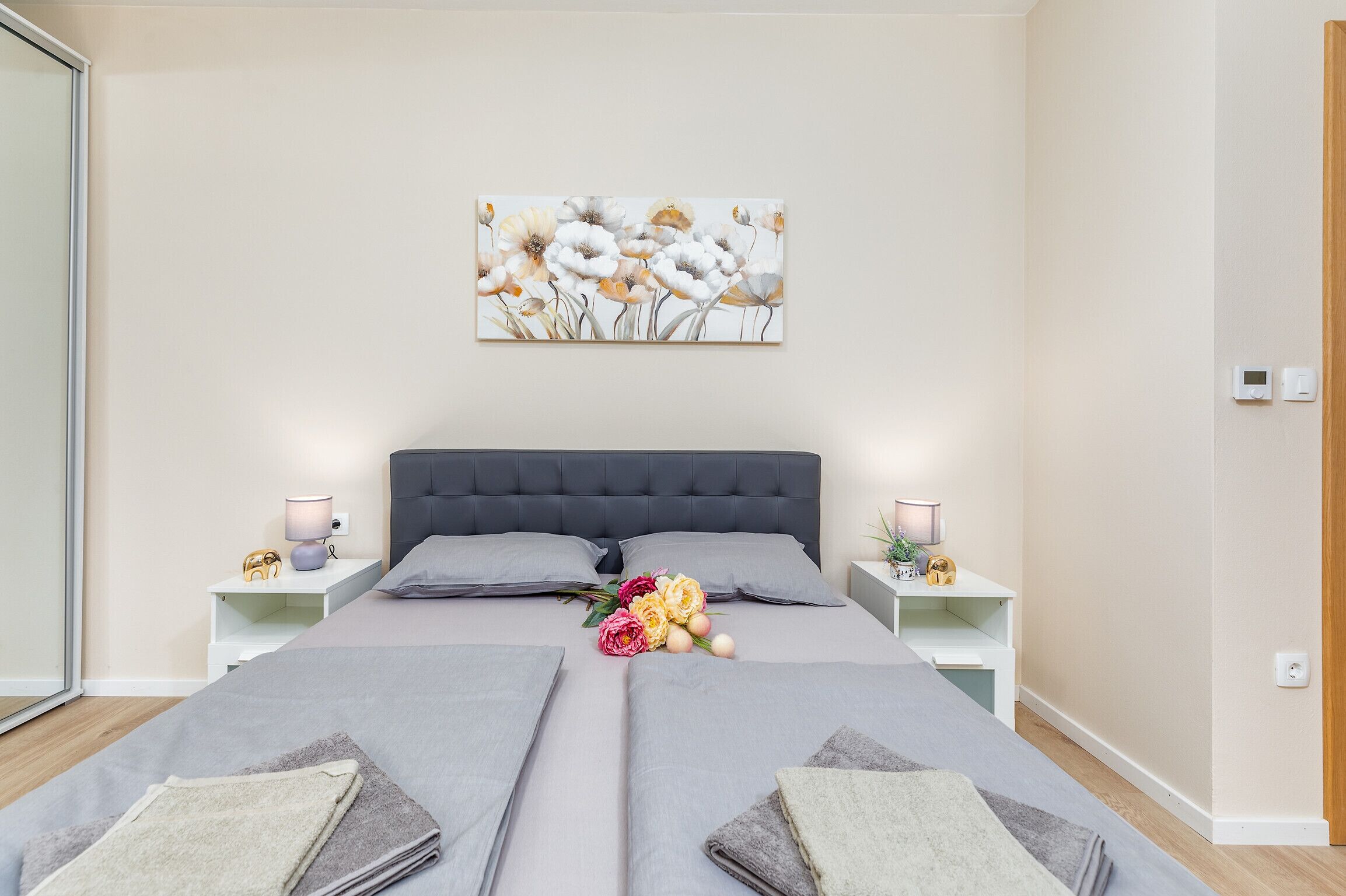 A modern bedroom with a large bed, two bedside tables, a nightstand, and a wall-mounted artwork.