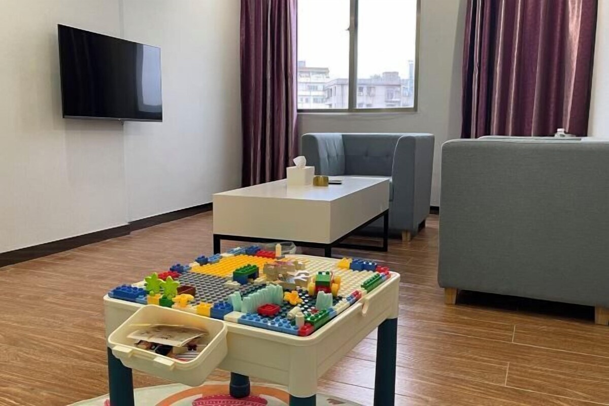Un salon équipé d’un téléviseur à écran plat, d’une petite table sur laquelle est posé un jeu de construction, d’un canapé gris et d’une fenêtre avec des rideaux.