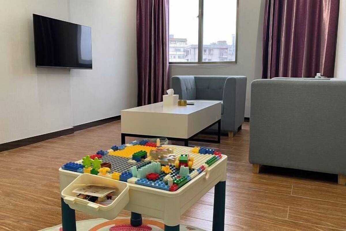 Una sala de estar con una televisión de pantalla plana, una mesita con un juego de juguetes, un sofá gris y una ventana con cortinas.