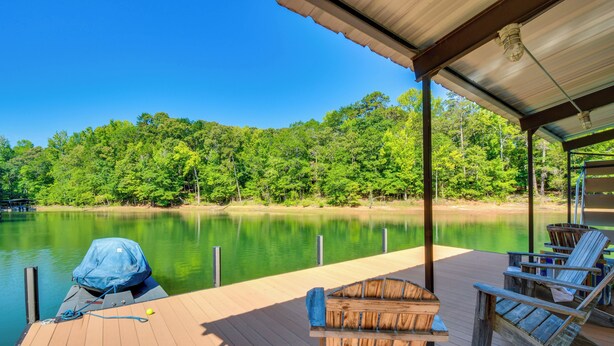 A wooden deck with a covered area, a boat, and chairs by a calm lake with a forest backdrop. - Dock Access & Patio! Waterfront Apt in Hartwell (Hartwell)