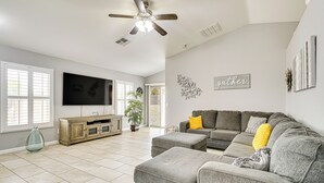 A spacious living room with a grey sectional sofa, a ceiling fan, a flat-screen TV, and a large window with blinds. - Tucson Home w/ Patio, Grill & Mountain Views! (Tucson)