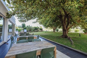 An outdoor dining area with wooden tables and green chairs, surrounded by a well-maintained lawn and a large tree providing shade. - T2 com Piscina Vilamoura Marina (Quarteira)