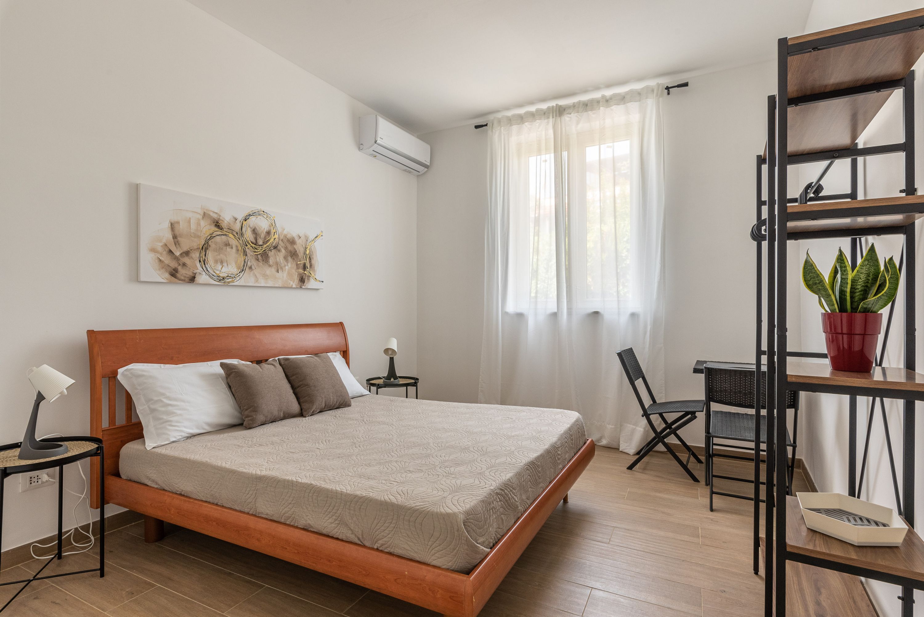 Quarto de hotel com cama de madeira, uma mesinha com abajur, uma cadeira, planta em vaso e prateleira na parede.