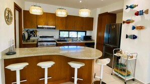 A modern kitchen with a central island, bar stools, wooden cabinets, stainless steel appliances, pendant lights, and decorative fish wall art. - Cozy 3 Bedroom Penthouse in Playa Bonita (Las Terrenas)