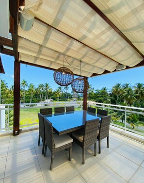 Uma área de refeições ao ar livre coberta, com mesa e cadeiras, com vista para uma paisagem exuberante com palmeiras.