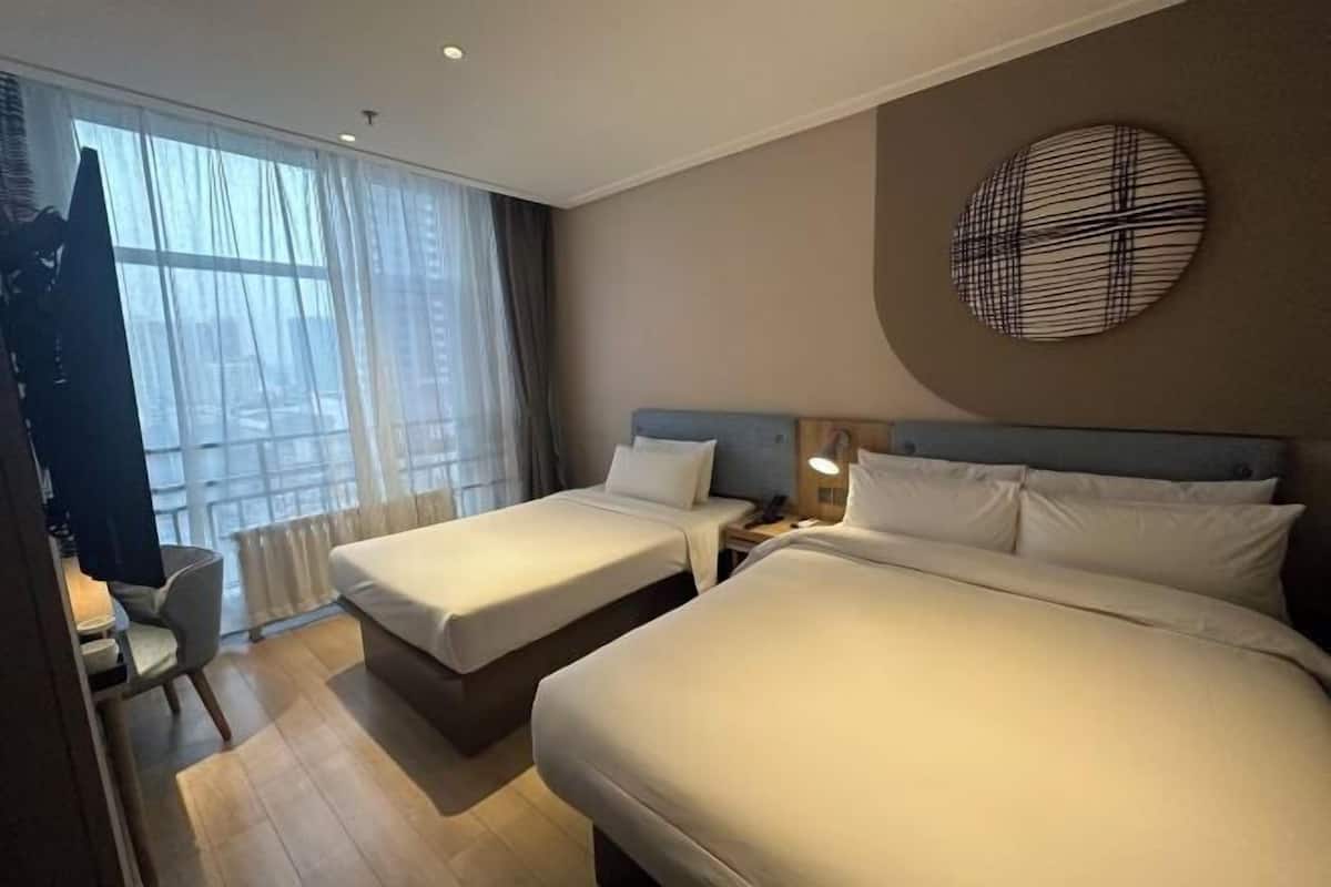 Et hotelværelse med to senge, et skrivebord, en stol og et stort vindue med gardiner.