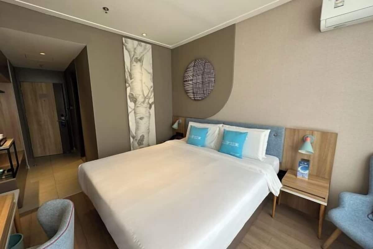 Et hotelværelse med en stor seng, et skrivebord, en stol, et natbord og en lampe på væggen.
