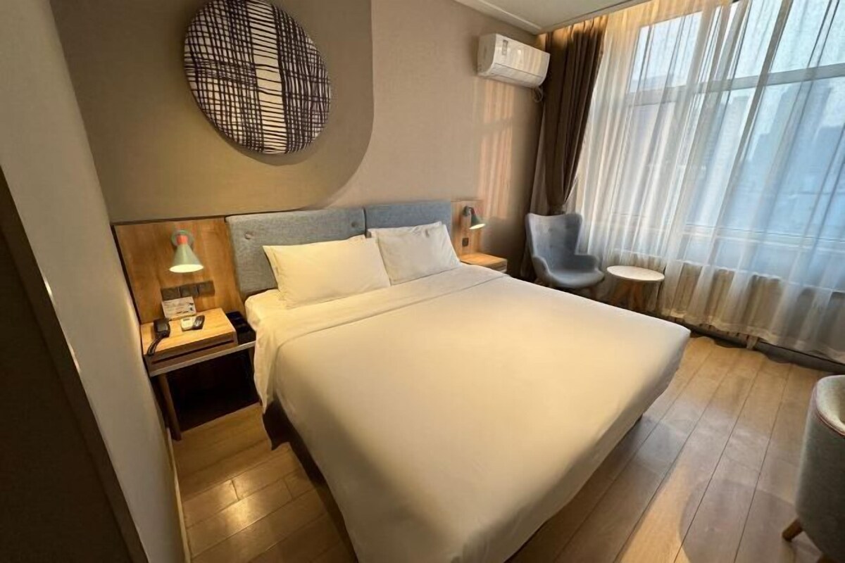 Ein Hotelzimmer mit einem großen Bett, einem Nachttisch mit Lampe, einem Sessel und einem Fenster mit durchsichtigen Vorhängen.