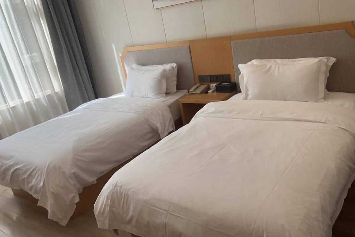 Ein Hotelzimmer mit zwei Einzelbetten, einem hölzernen Kopfteil, einem Nachttisch mit Telefon und einem Fenster mit leichtem Vorhang.
