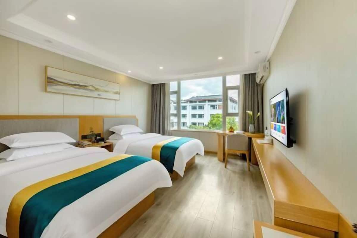 Ein Hotelzimmer mit zwei Betten, einem Schreibtisch, einem Fernseher und einem Fenster mit Blick auf Gebäude.