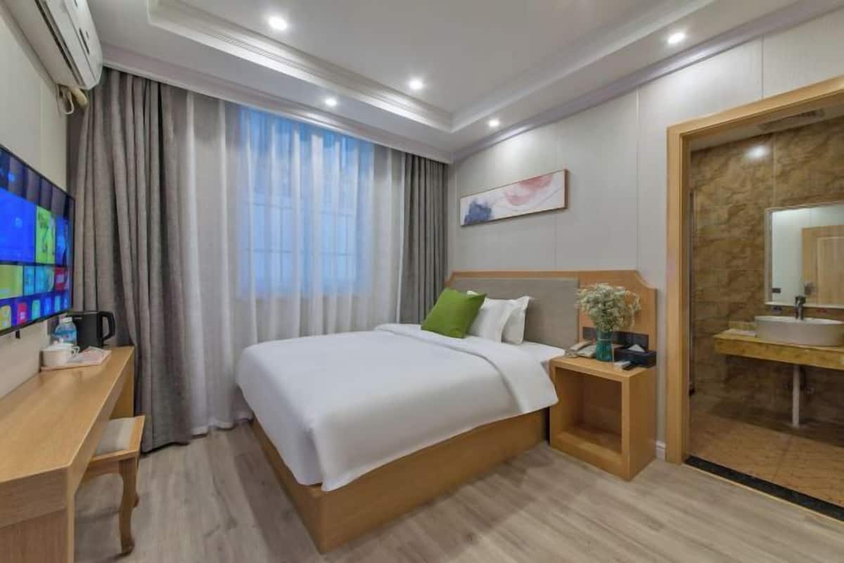 Ein Hotelzimmer mit Bett, Schreibtisch, Stuhl, Fernseher, einem Fenster mit Vorhängen, einem Gemälde und einem Badezimmer mit Waschbecken und Spiegel.
