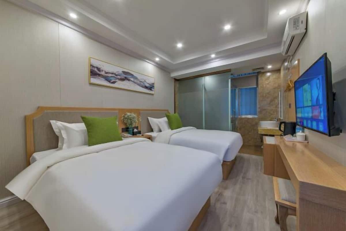 Ein Hotelzimmer mit zwei Betten, einem Schreibtisch, einem Fernseher und einem durch eine Glaswand abgetrennten Badezimmer.