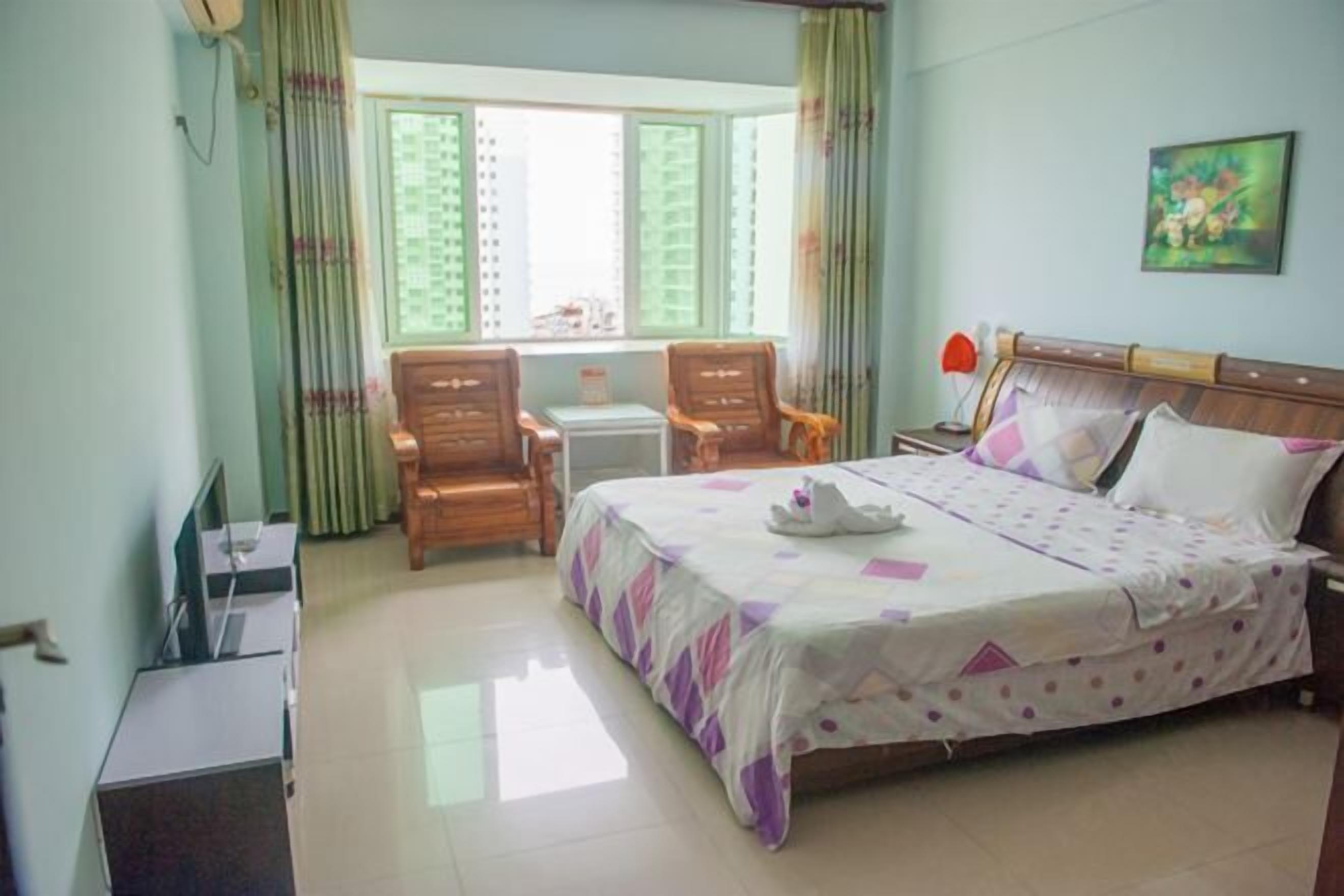 A hotel room with a bed, two wooden chairs, a small table, a TV, and a window with curtains.