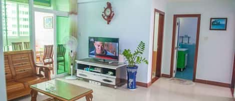 Uma sala de estar com televisão, móveis de madeira, um vaso com planta e uma porta de vidro deslizante para uma varanda.
