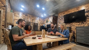 Een groep mensen speelt een kaartspel aan een houten tafel in een kamer met een houten inrichting en een aan de muur bevestigde flatscreen televisie.