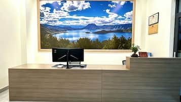 Un mostrador de recepción con un monitor de computadora, un cuadro enmarcado de un paisaje encima y una pequeña planta en maceta a la derecha.
