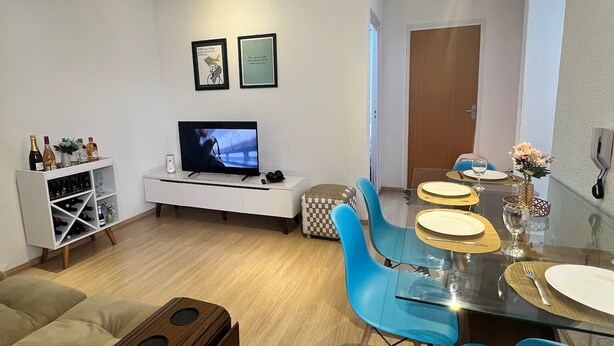A modern living room with a flat-screen TV, a white entertainment unit, and a dining area with blue chairs and a glass table.