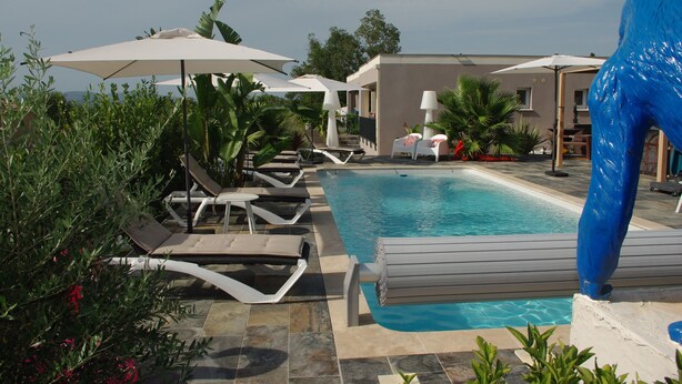 Un espace piscine avec des chaises longues et des parasols, entouré de verdure et avec un bâtiment en arrière-plan.