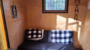 A cozy room with a wooden wall, a window with a view of trees, a checkered sofa, a patterned rug, and a small heater. - The Overleigh Shearers Quarters (Port Levy)