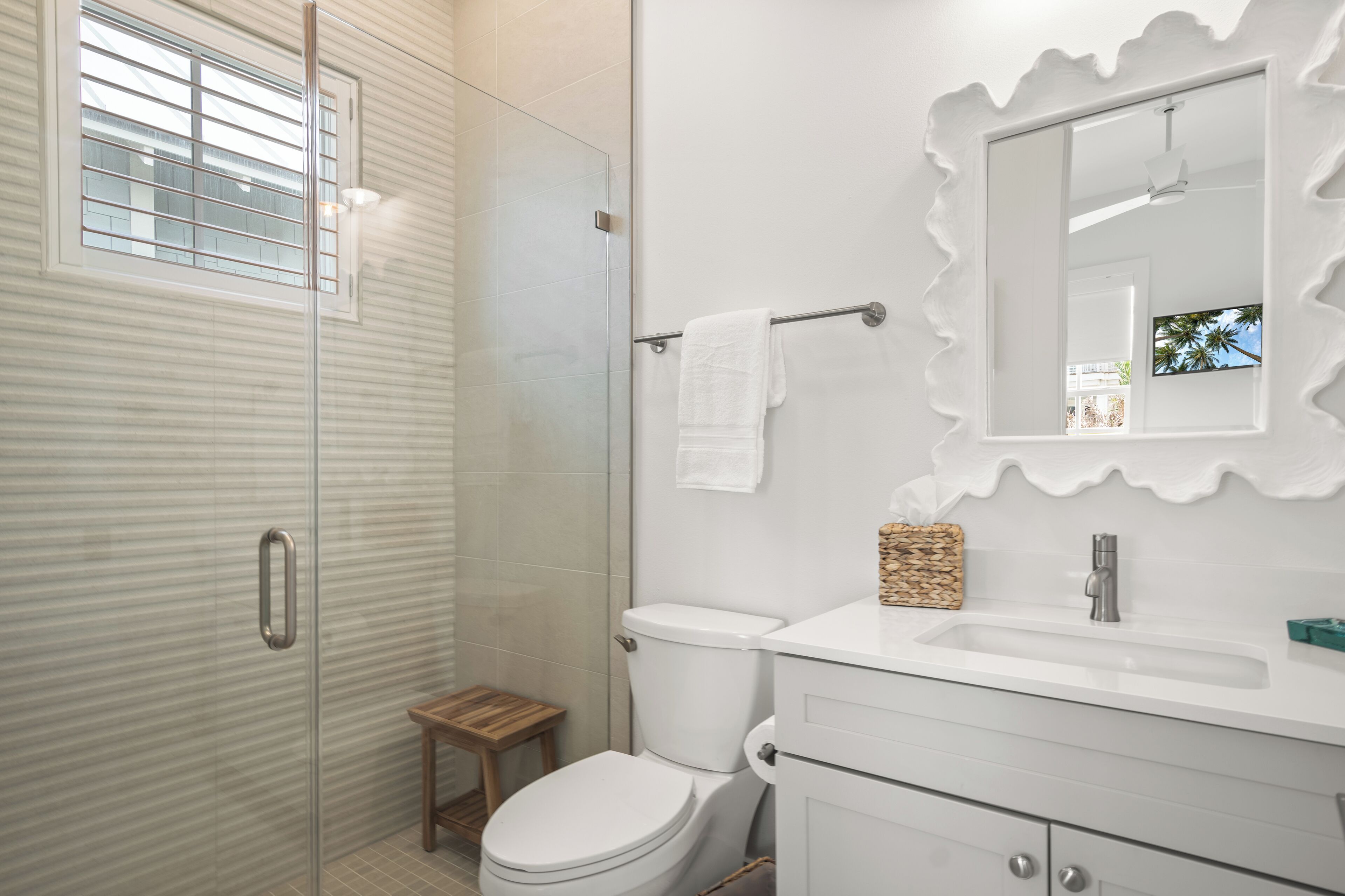 A bathroom with a glass shower enclosure, a white toilet, a vanity with a mirror, and a small wooden stool.