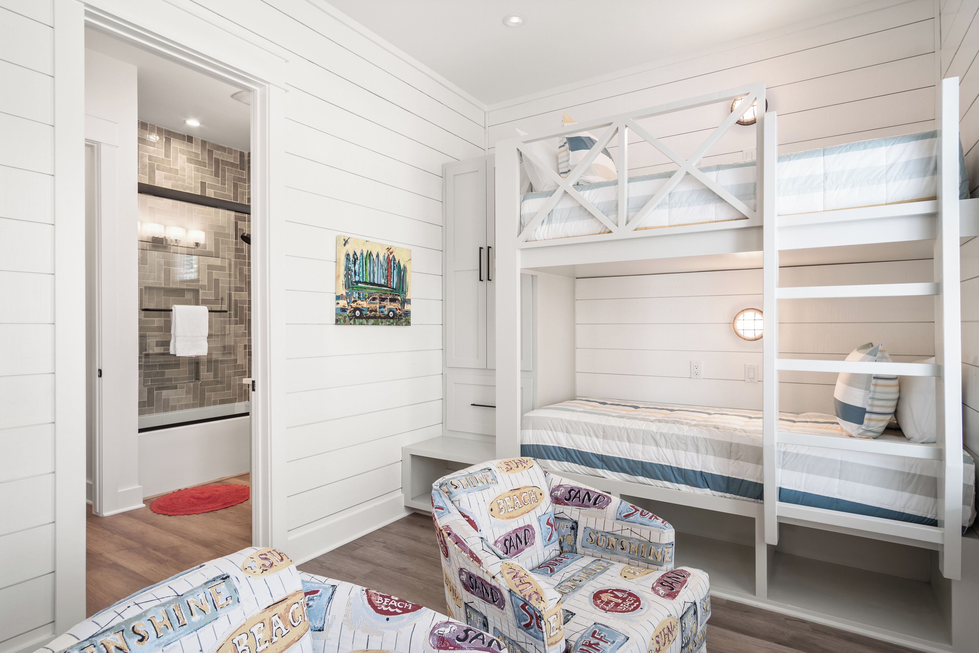 A bedroom with bunk beds, a chair with beach-themed upholstery, and a bathroom with a tiled shower wall.