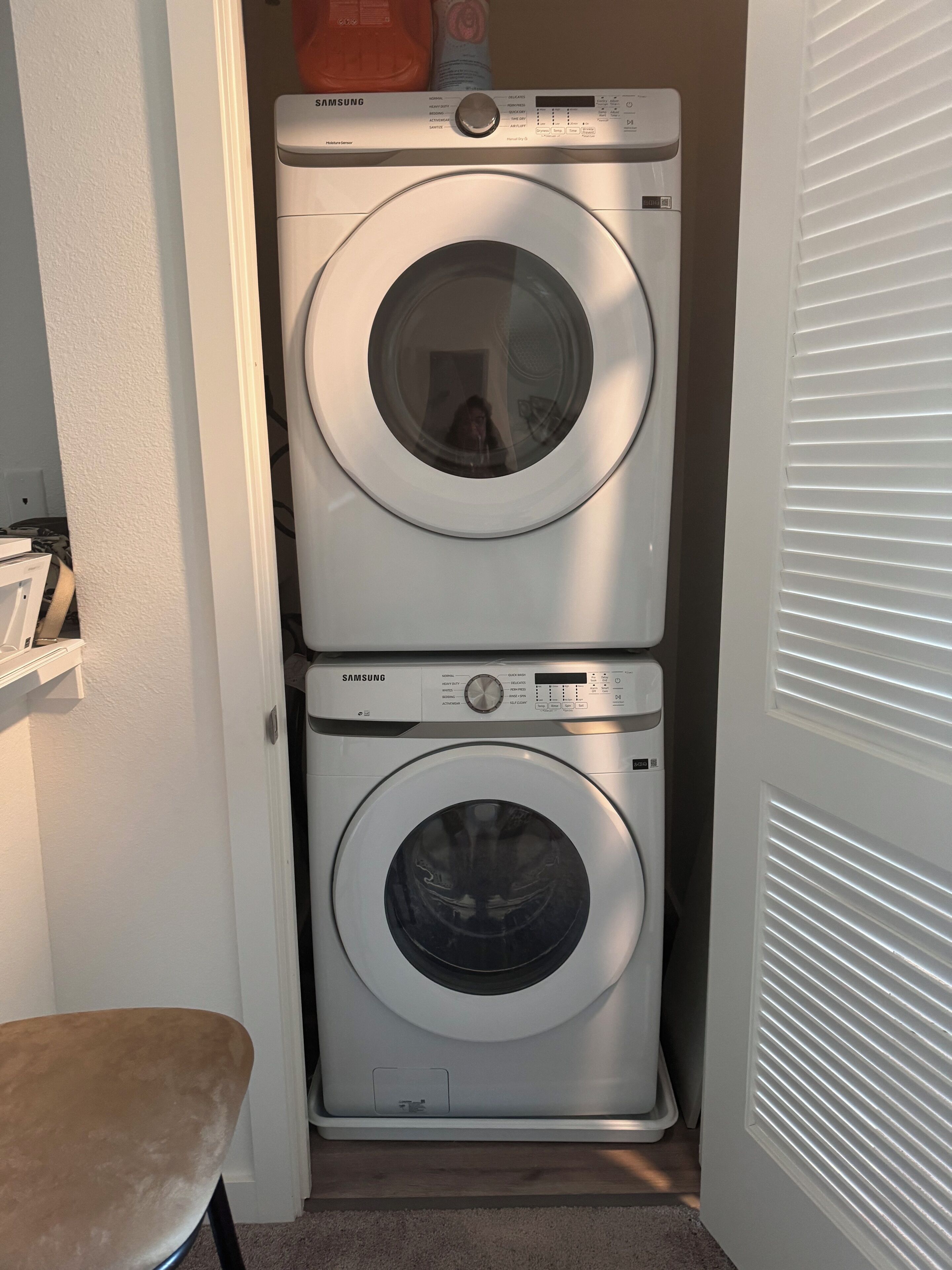 A Samsung front-loading washer and dryer stacked in a small laundry room with a wooden bench and a door with horizontal blinds.