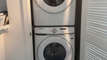 A Samsung front-loading washer and dryer stacked in a small laundry room with a wooden bench and a door with horizontal blinds.
