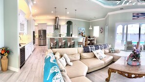A spacious living room with a large sofa, a coffee table, and a TV. There is a kitchen area with white cabinets and a dining area with a table and chairs.